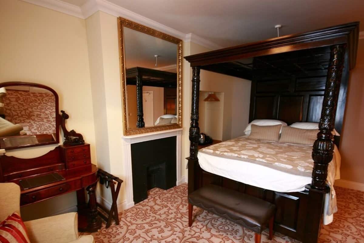 A four-poster bed with a white bedspread, a wooden bench at the foot, a large mirror with a gold frame, and a patterned carpet.
