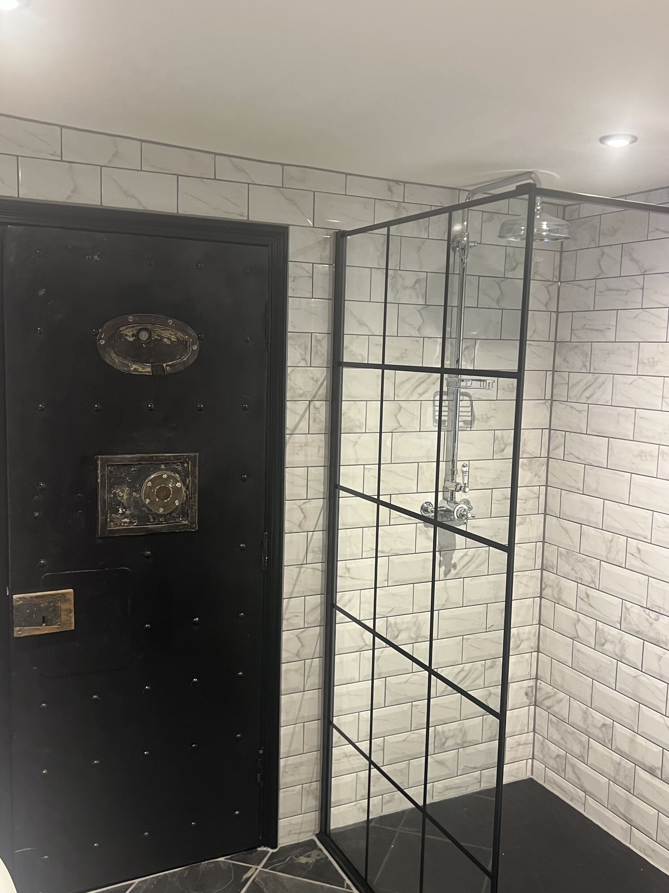 A black door with a metal handle and a small plate, a glass-enclosed shower with a metal frame, and white marble tiles on the walls.