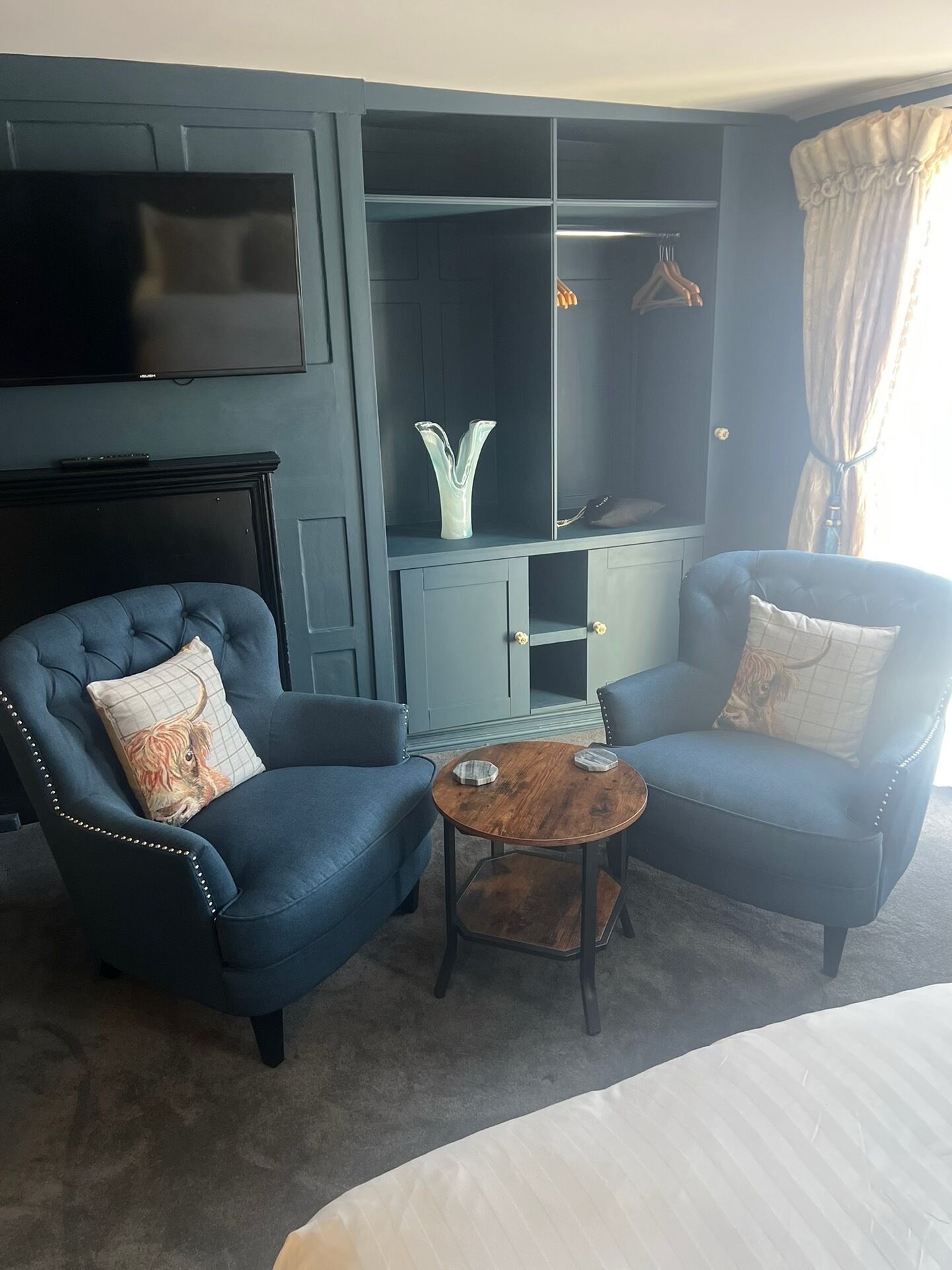 A modern living room with two blue armchairs, a small wooden coffee table, a TV mounted on the wall, and a grey cabinet with a decorative vase.