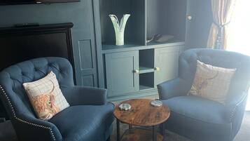 A modern living room with two blue armchairs, a small wooden coffee table, a TV mounted on the wall, and a grey cabinet with a decorative vase.