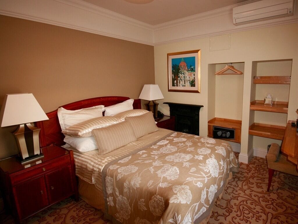 A hotel room with a large bed, two bedside tables with lamps, a dresser, a fireplace, a framed picture on the wall, and a wooden chair.