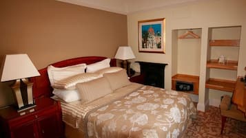 A hotel room with a large bed, two bedside tables with lamps, a dresser, a fireplace, a framed picture on the wall, and a wooden chair.