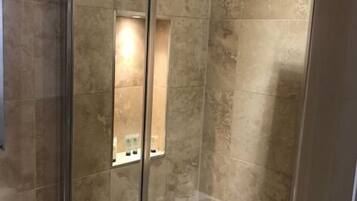 Ducha con cerramiento de vidrio, cabezal de ducha de mano, puerta de cristal y un pequeño estante con artículos de aseo.