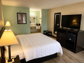 A hotel room with a bed, a desk with a lamp, a television, a dresser, and a bathroom visible in the background.