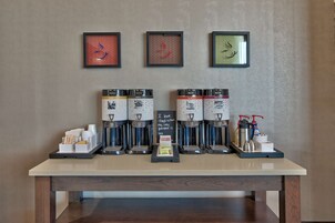 Una estación de café con varias máquinas de café, una bandeja con tazas y un cartel, y obras de arte enmarcadas en la pared.