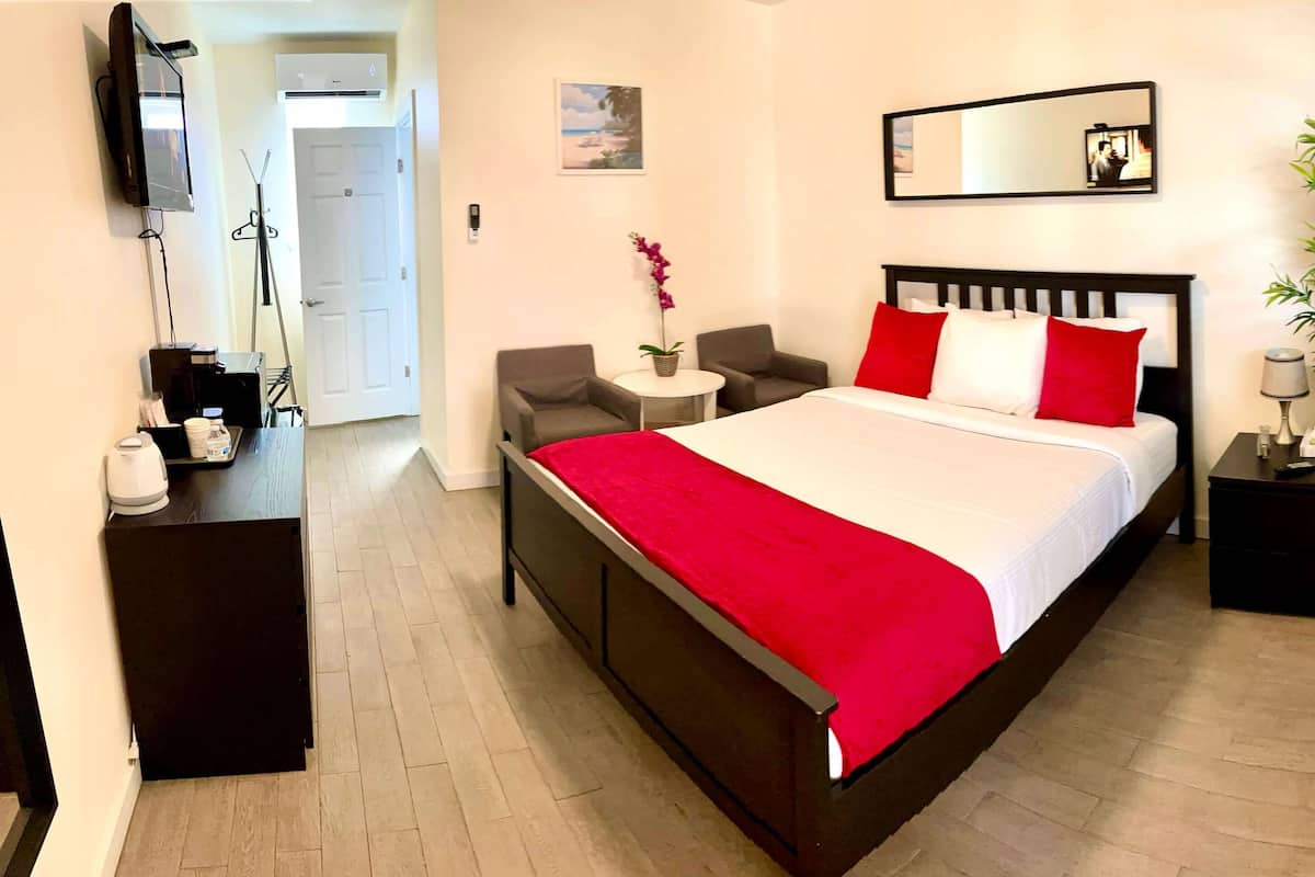 A hotel room with a bed, bedside tables, a TV, a desk, a chair, a potted plant, and a door.