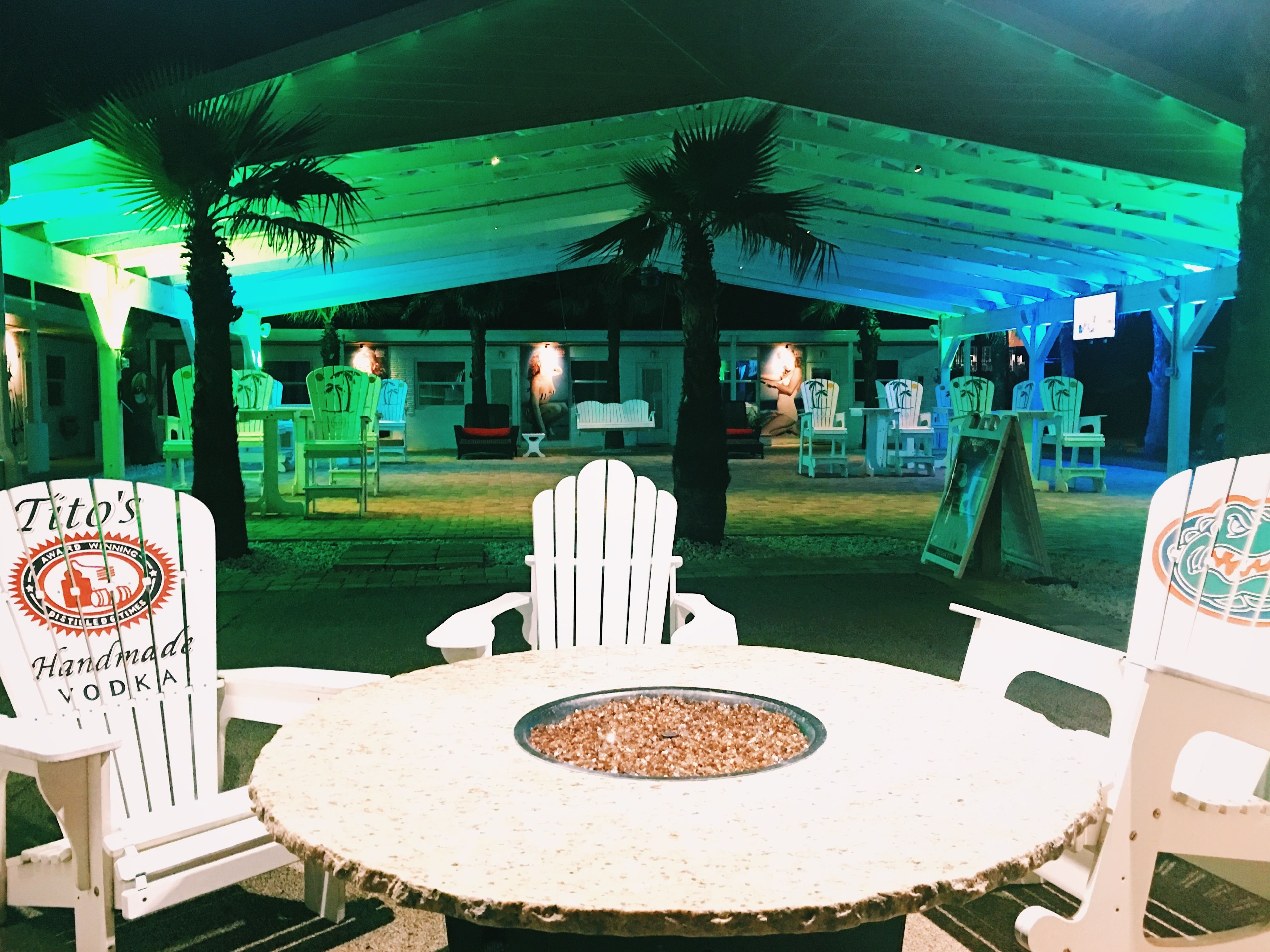 A covered outdoor seating area with white Adirondack chairs and a round table with a fire pit.