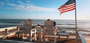 Duas cadeiras Adirondack num deck de madeira com vista para o oceano e uma bandeira americana.