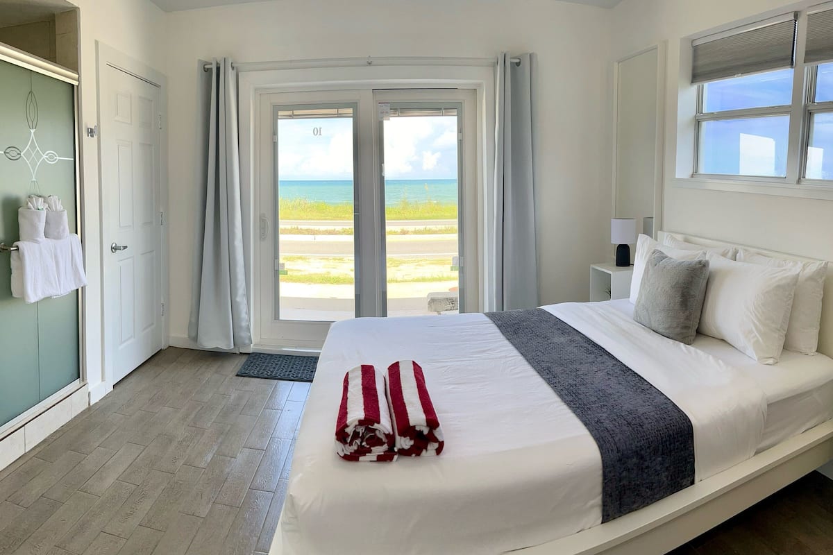 A hotel room with a bed, a nightstand, a lamp, a potted plant, and a view of the beach.
