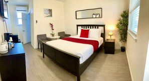 Quarto de hotel com cama, mesas de cabeceira, escrivaninha com chaleira e garrafa de água, pequena mesa redonda com planta e janela com persianas.
