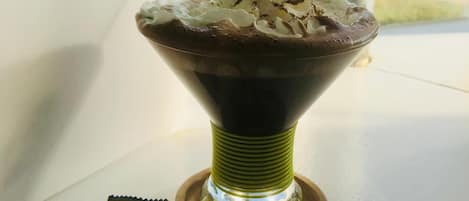 Un dessert servi dans un bol en verre, garni de crème fouettée et de morceaux de chocolat, est posé sur une table près d’une fenêtre.