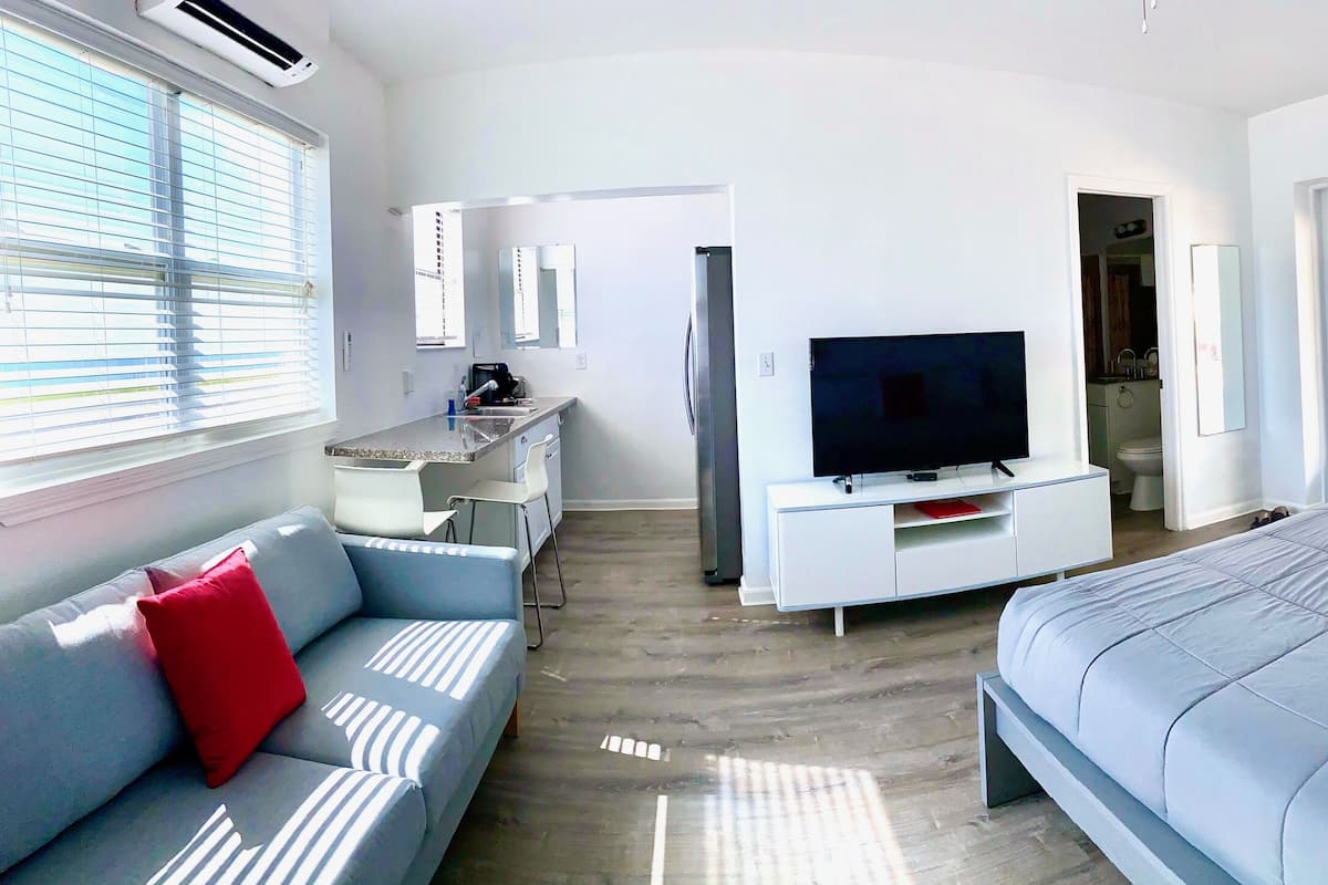 A modern hotel room with a large bed, a TV, a small kitchenette, and a seating area.