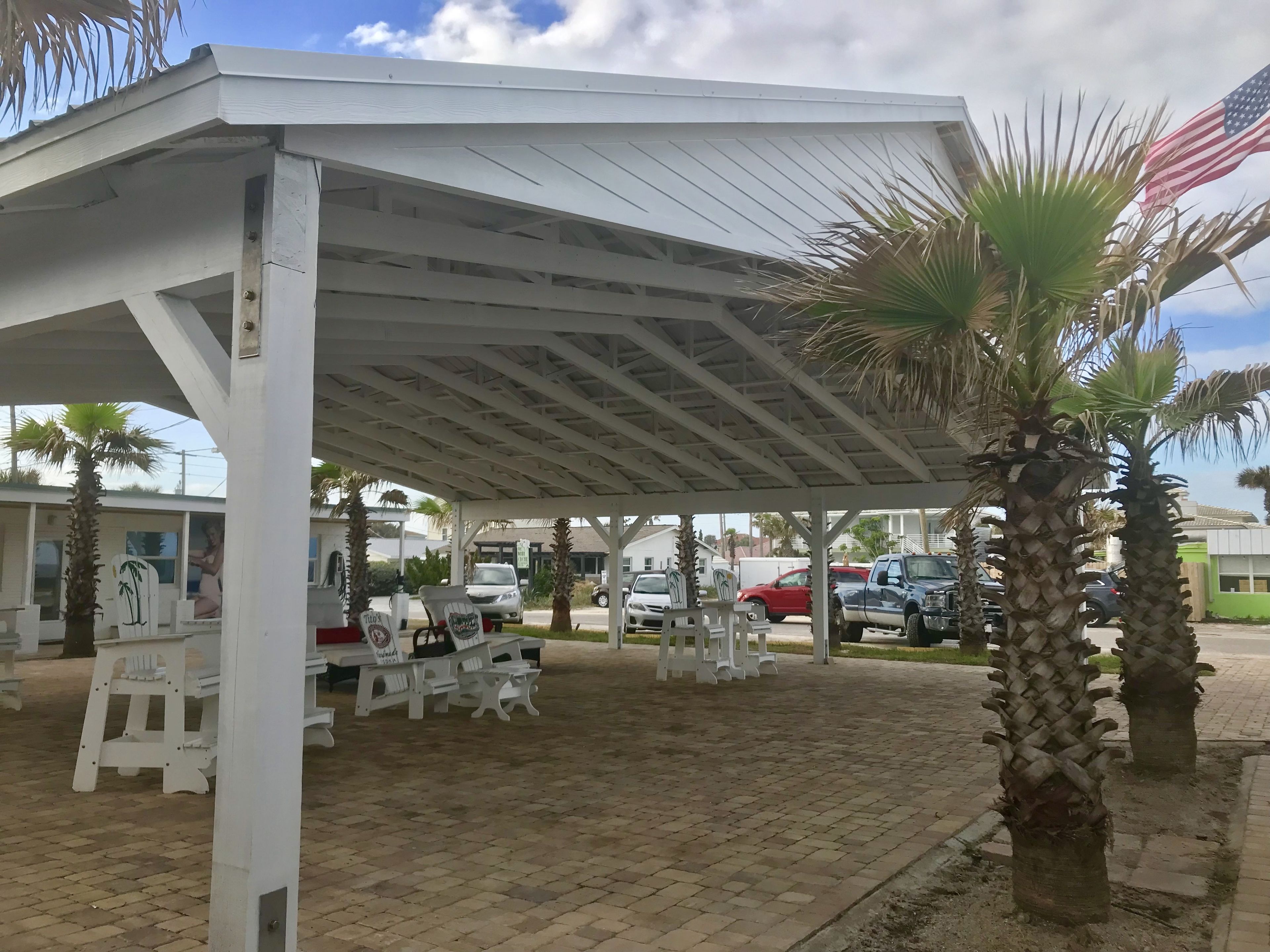 A covered outdoor seating area with white benches and tables, palm trees, and a view of parked cars and buildings in the background.