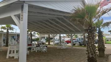 A covered outdoor seating area with white benches and tables, palm trees, and a view of parked cars and buildings in the background.