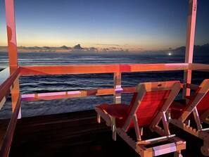 Um deck de madeira com duas cadeiras voltadas para a vista do oceano ao pôr do sol.