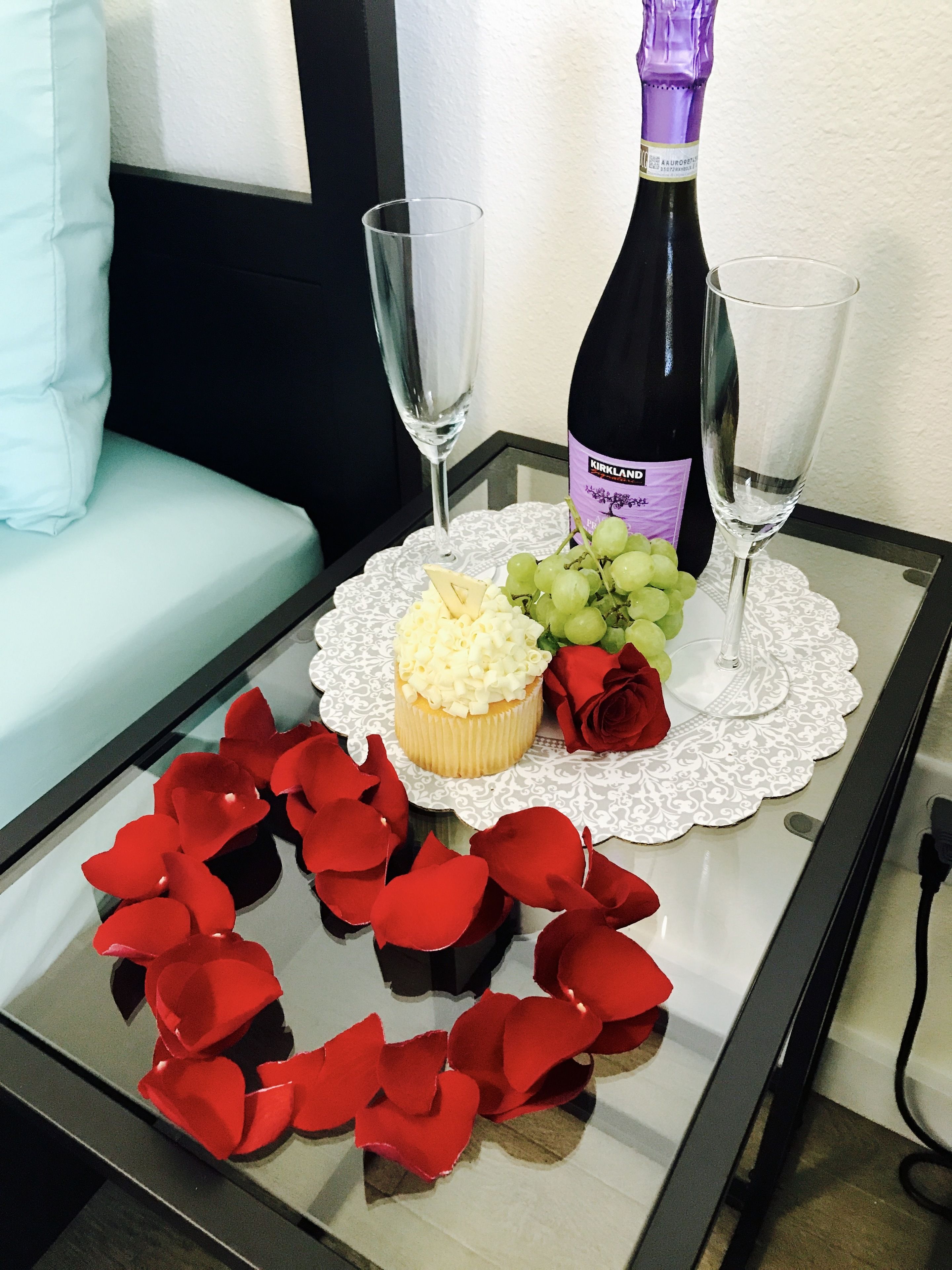 Una mesa de vidrio con un posavasos de encaje blanco, una botella de vino Kirkland, dos copas de vino, un cupcake, un racimo de uvas y pétalos de rosa roja.