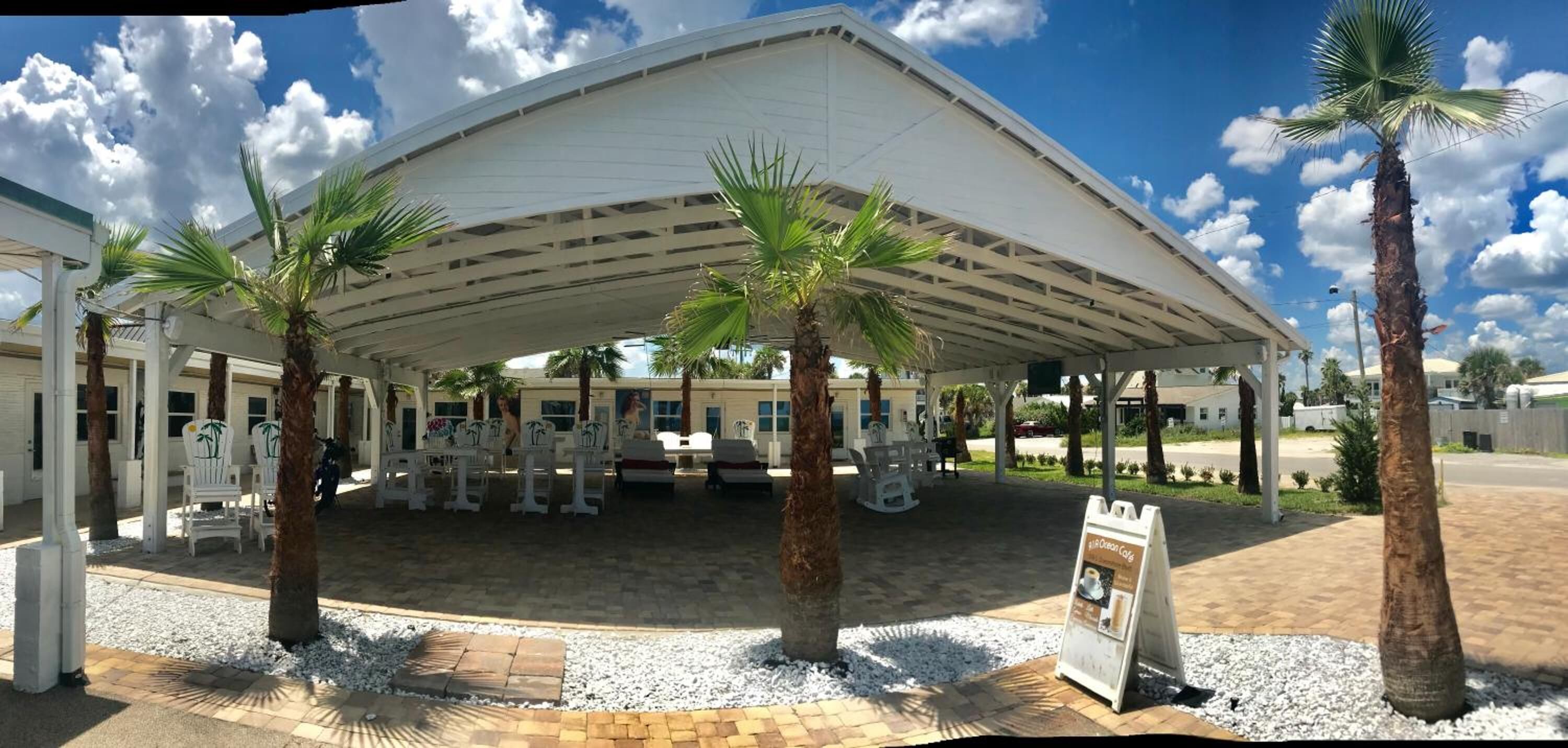 A covered outdoor seating area with palm trees, a paved walkway, and a signboard.