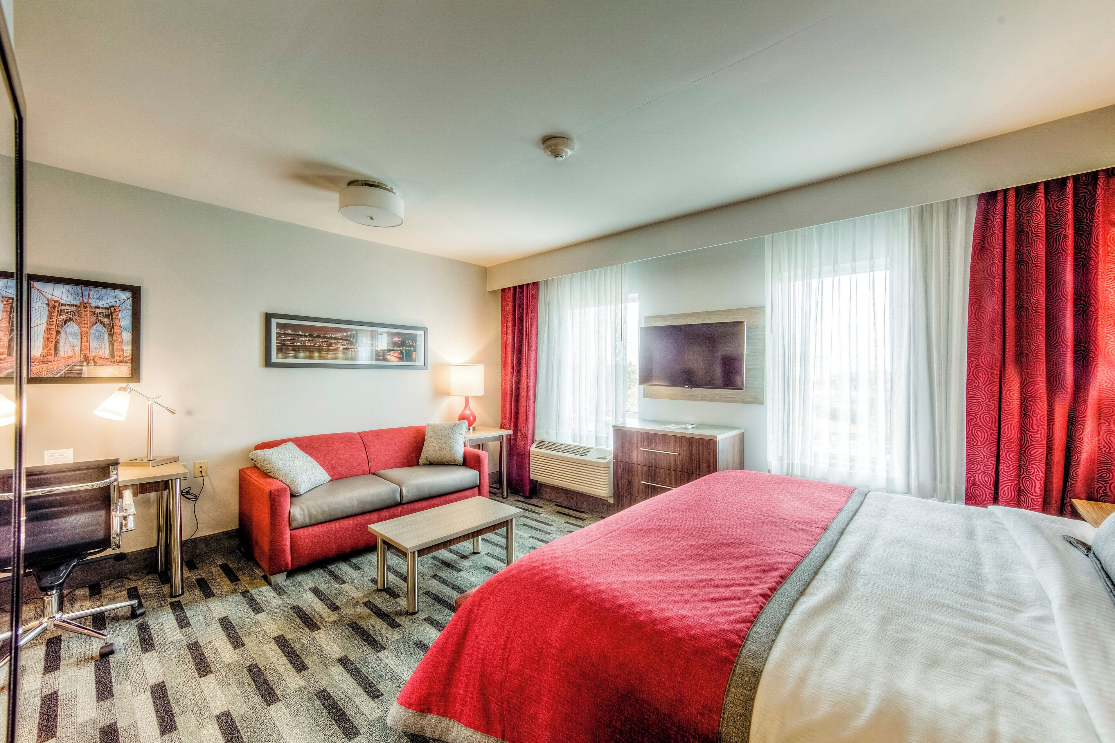 A hotel room with a bed, a sofa, a desk, a lamp, a TV, and a window with curtains.
