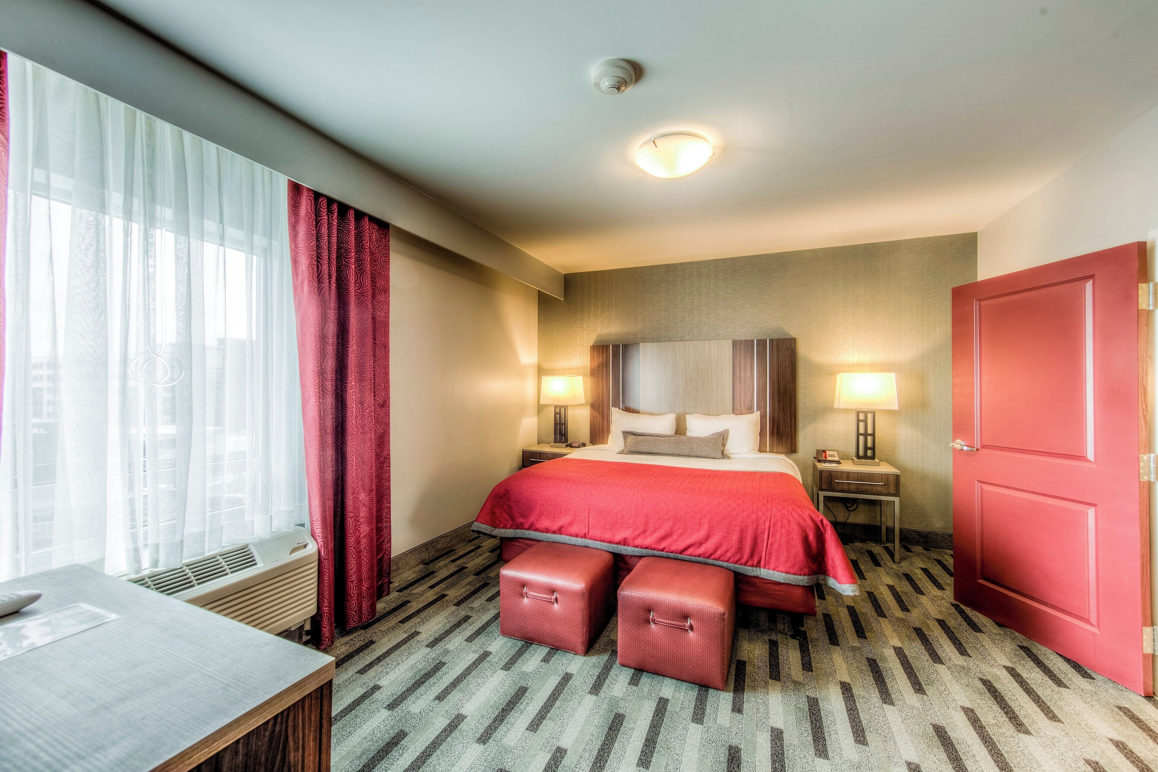 Quarto de hotel com cama, mesas de cabeceira, escrivaninha e janela com cortinas.