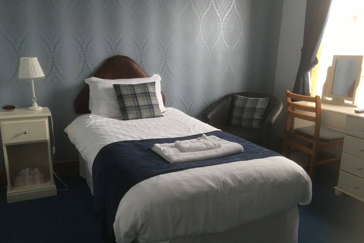 A neatly made bed with a blue blanket, a white pillow, and a plaid pillow. There is a nightstand with a lamp and a small table with a tissue box. A wooden chair and a desk with a computer are visible.