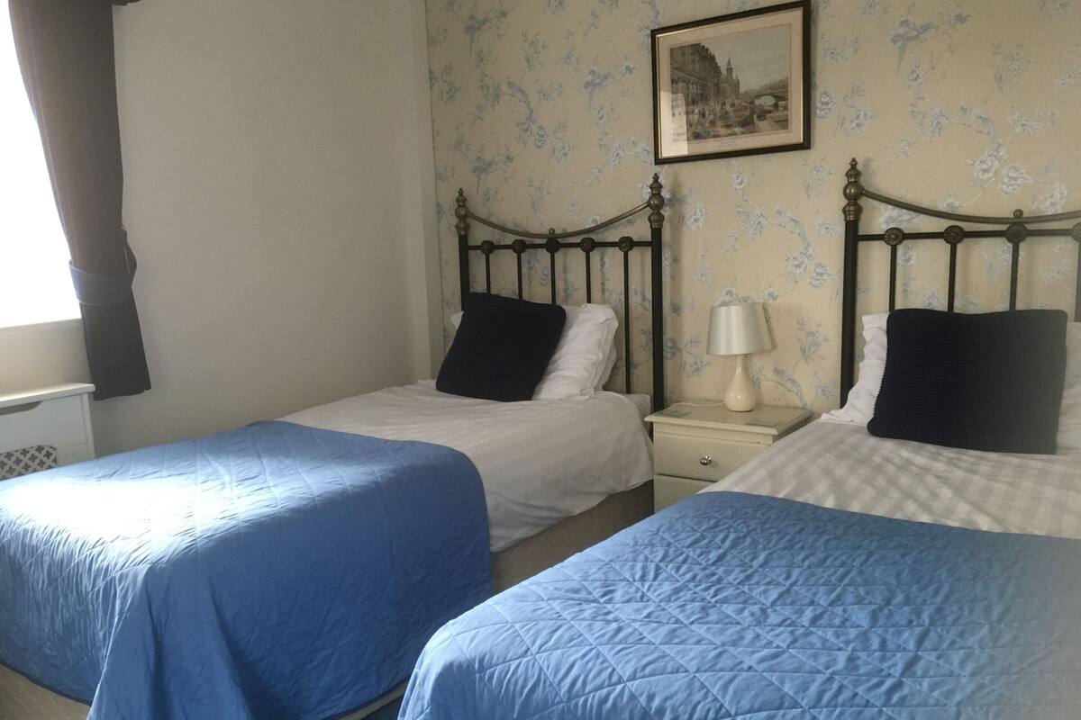 A room with two beds, each with a blue bedspread and black pillows. There is a nightstand with a lamp between the beds. The walls have a floral wallpaper pattern, and a framed picture hangs on the wall.
