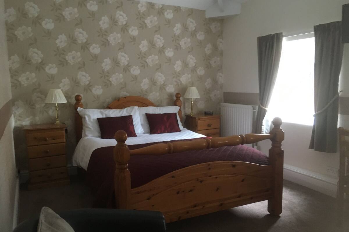 A bedroom with a wooden bed, two bedside tables, a nightstand, a radiator, and a window with curtains.