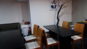 A small, dimly lit room with a dark sofa, a table, and four wooden chairs. A white cylindrical object, possibly a trash can, is against the wall.