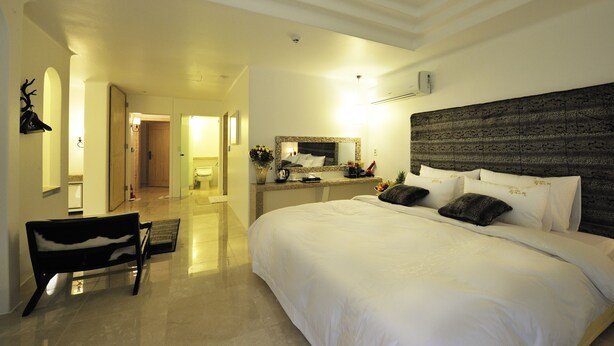 A hotel room with a large bed, a bench, a mirror, and a door leading to a bathroom. - MS Hotel (Suwon)