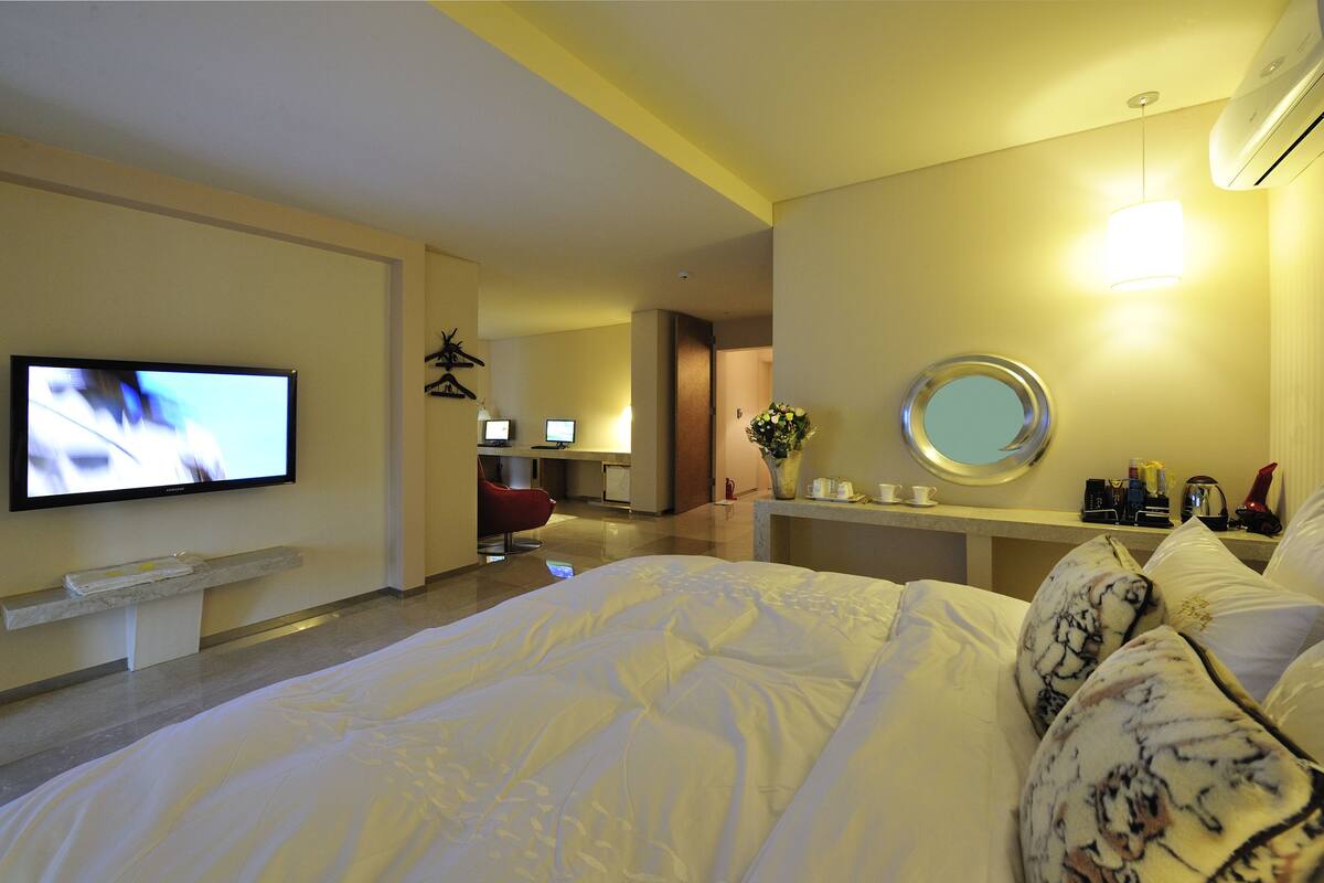 A hotel room with a bed, a TV, a desk with a chair, a mirror, and a vase with flowers.
