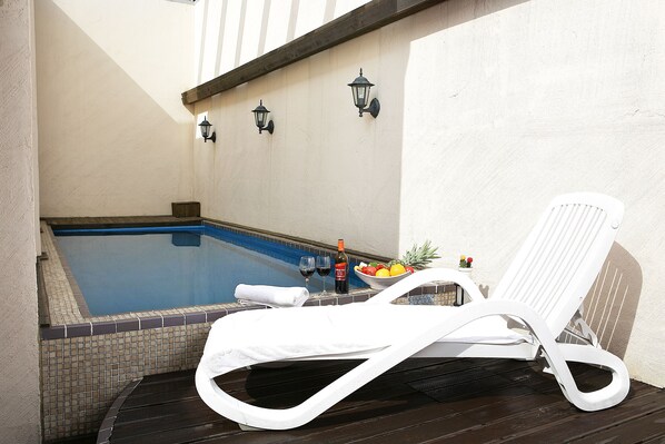 A sun lounger by a pool with a tiled edge, a bottle of wine, and a bowl of fruit.