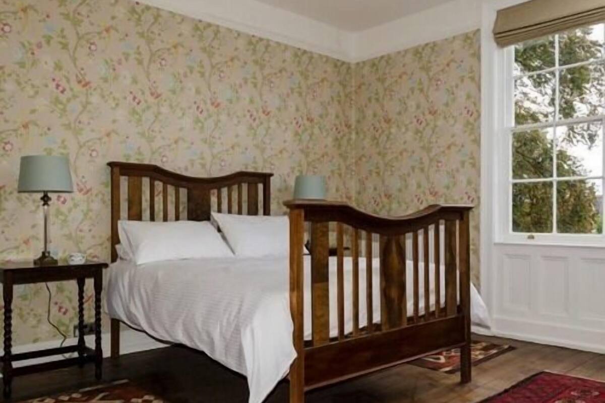 Quarto com cama de madeira, mesa de cabeceira, luminária e janela com vista para árvores.