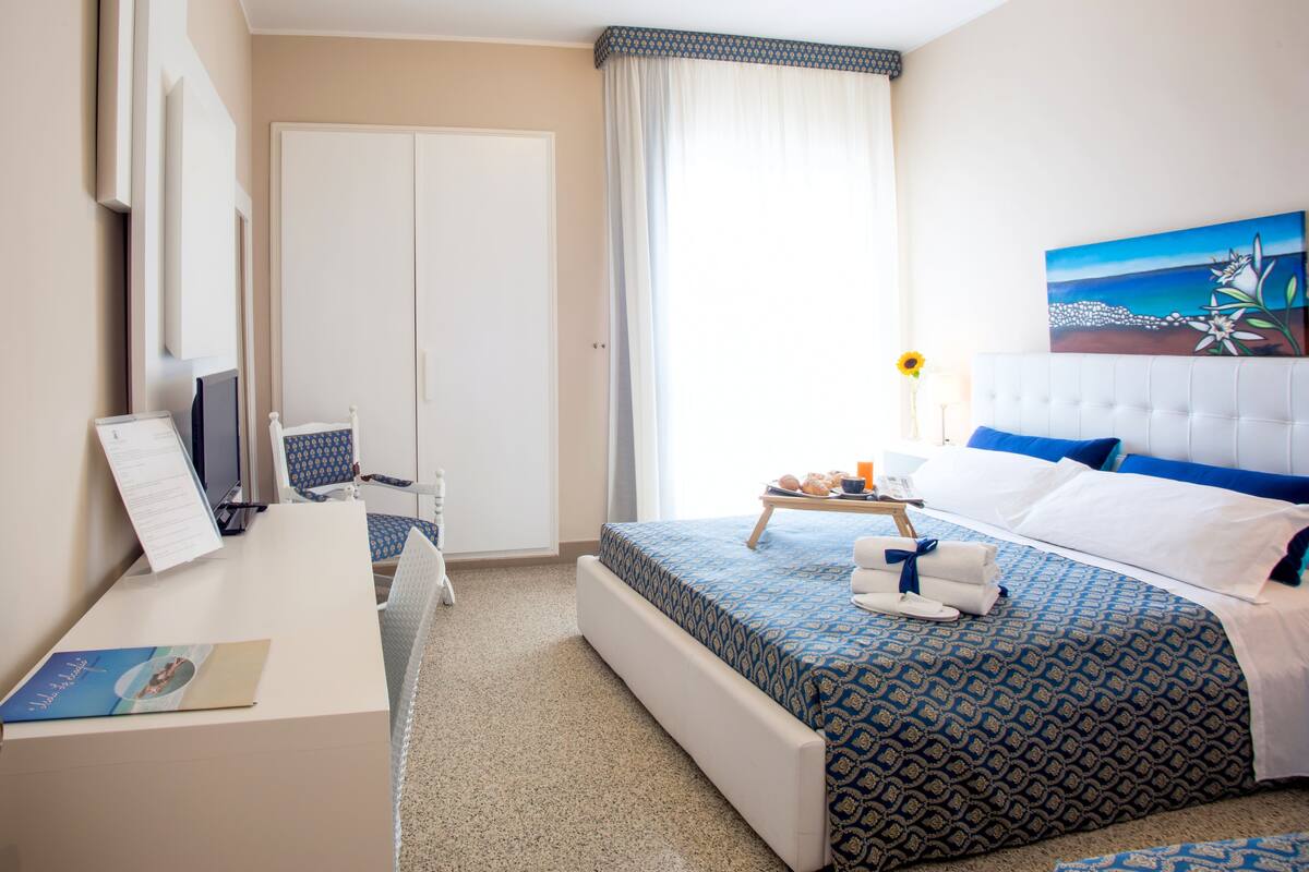 Habitació d'hotel amb llit, escriptori, cadira, tauleta de nit, quadre i finestra amb cortines.