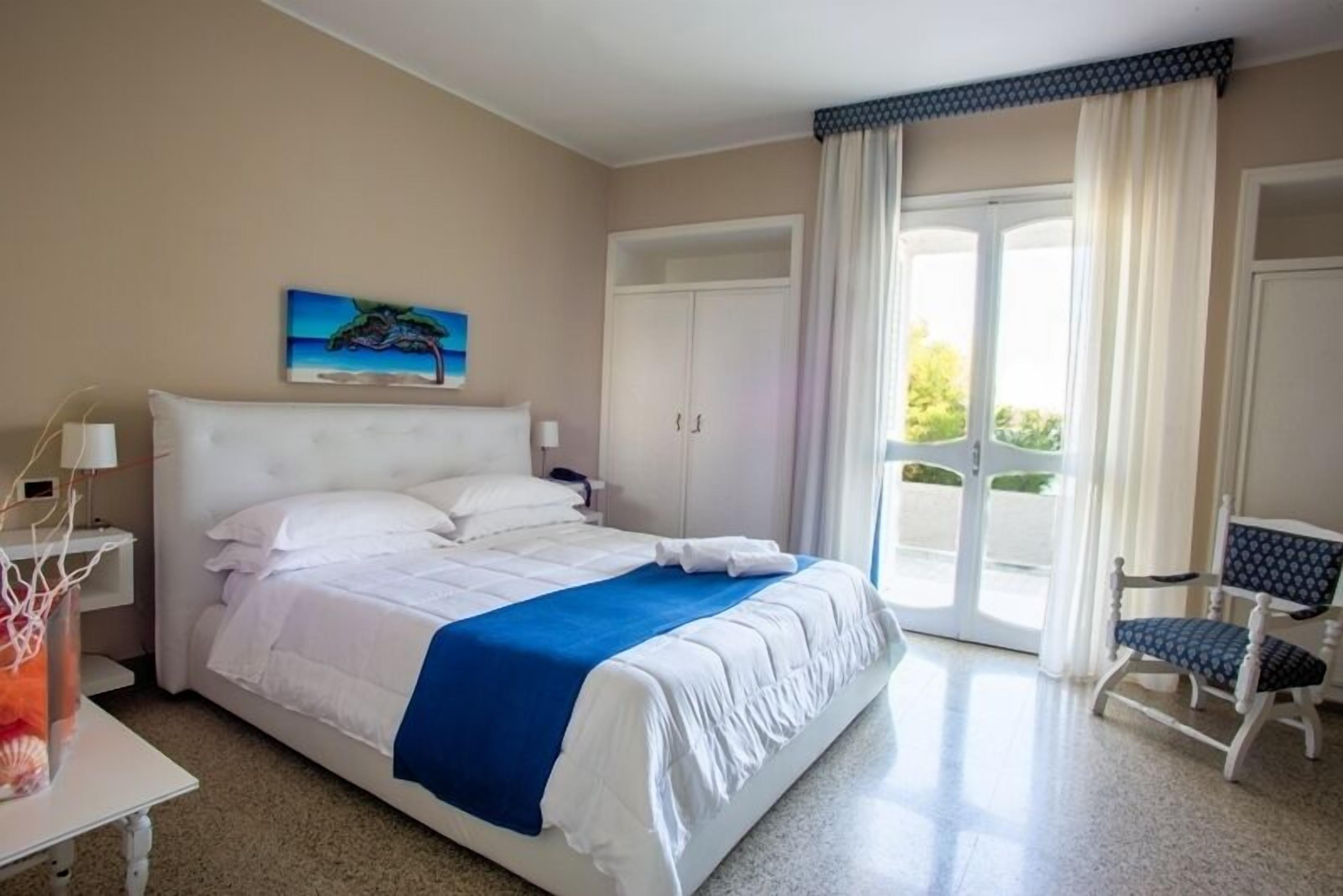 A hotel room with a large bed, a desk, a chair, a painting, and a sliding glass door to the outside.