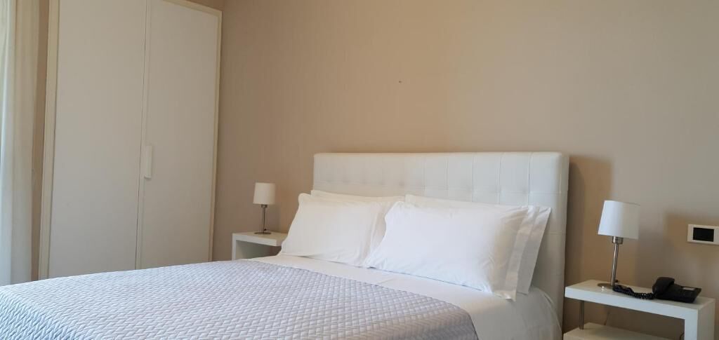 Un letto ben rifatto con lenzuola bianche, due comodini con lampade e una porta chiusa.