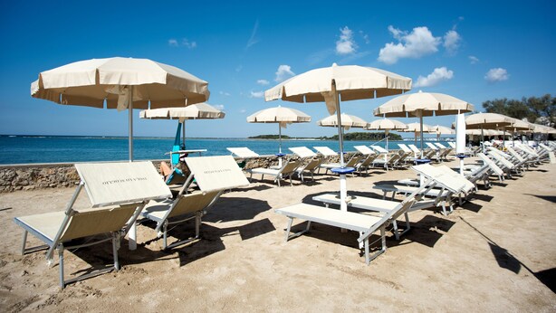 Ein Strand mit Reihen von Liegestühlen und weißen Sonnenschirmen, einem klaren blauen Himmel und ruhiger See.