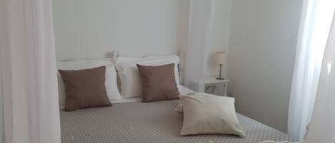 A neatly made bed with white and brown bedding, a small bedside table with a lamp, and sheer curtains.