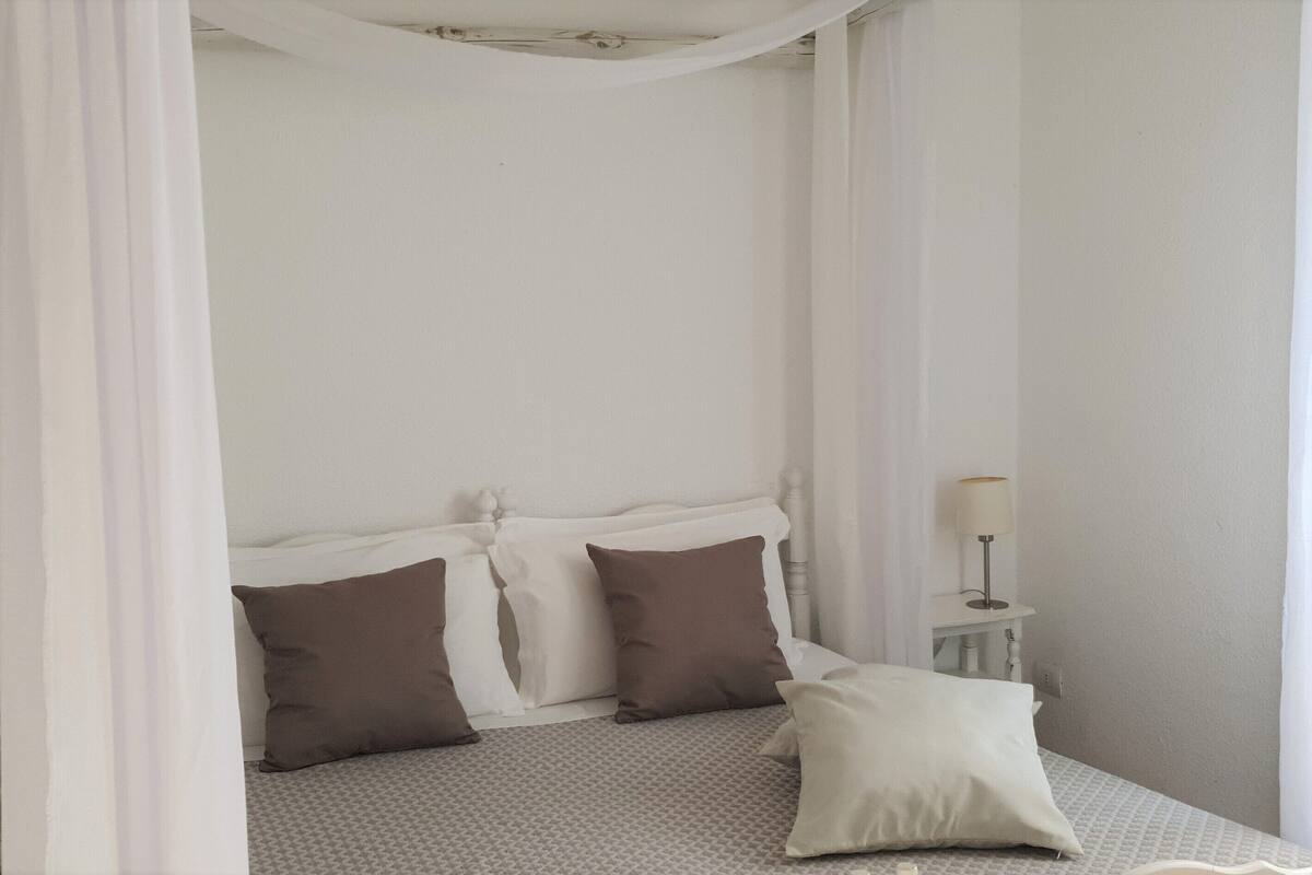 Un llit ben fet amb roba de llit blanca i marró, una tauleta de nit amb una làmpada i cortines translúcides.