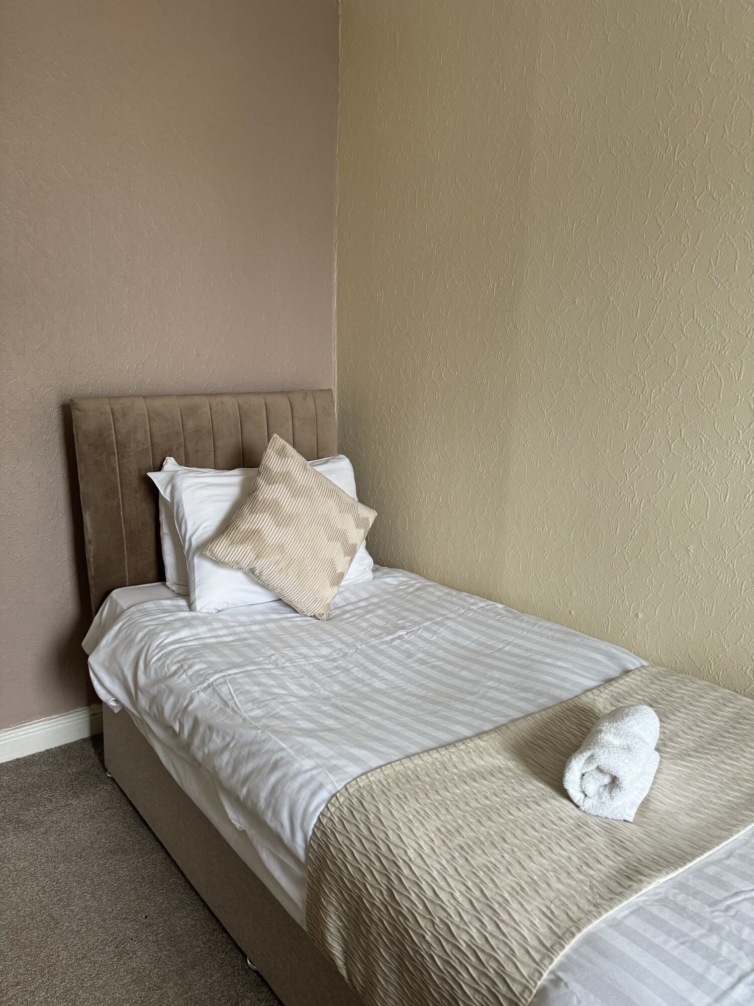 Una cama bien hecha con cabecera, una toalla enrollada y dos almohadas.