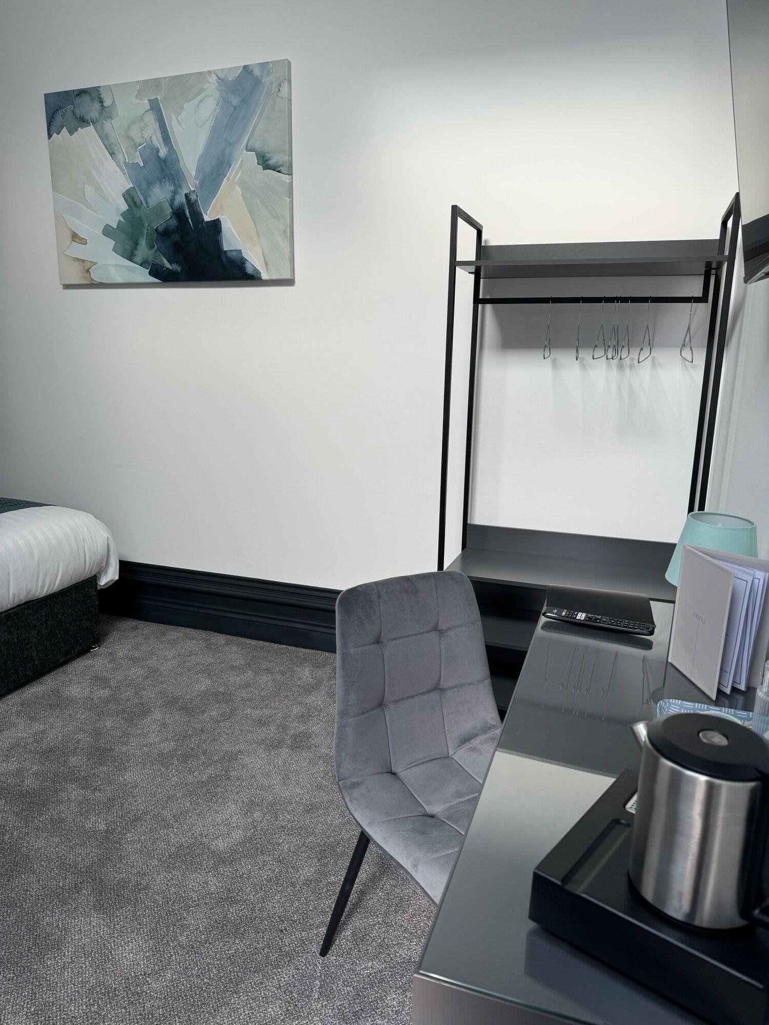 A modern hotel room with a bed, a gray upholstered chair, a desk with a kettle, and a painting on the wall.