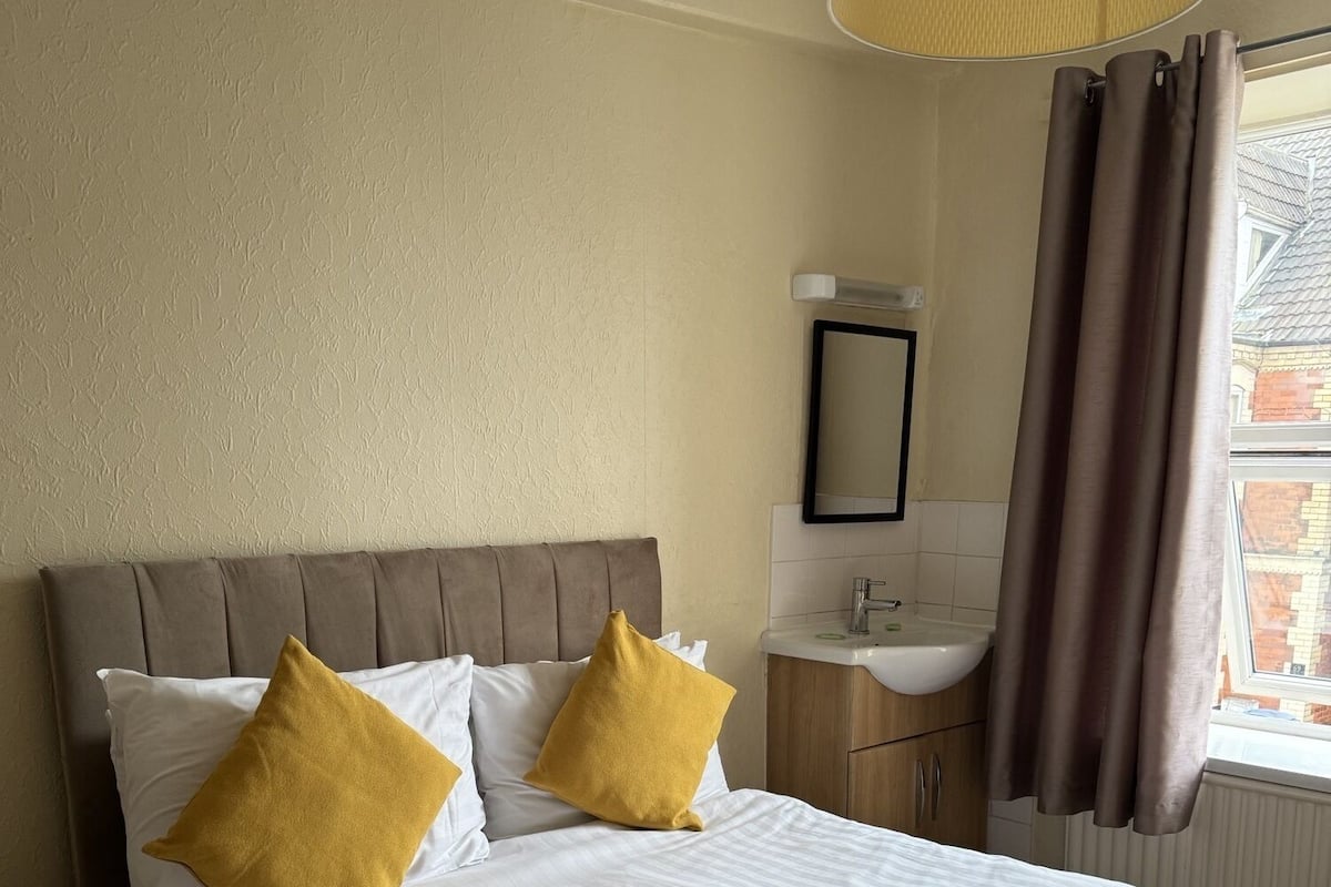 Quarto de hotel com cama, mesa de cabeceira, espelho, janela com cortinas e banheiro com pia.