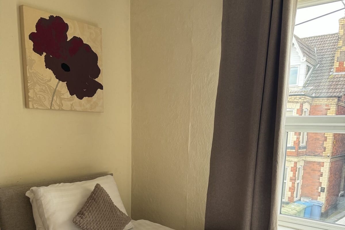 Quarto com cama, almofadas, manta, uma obra de arte na parede e janela com cortinas.