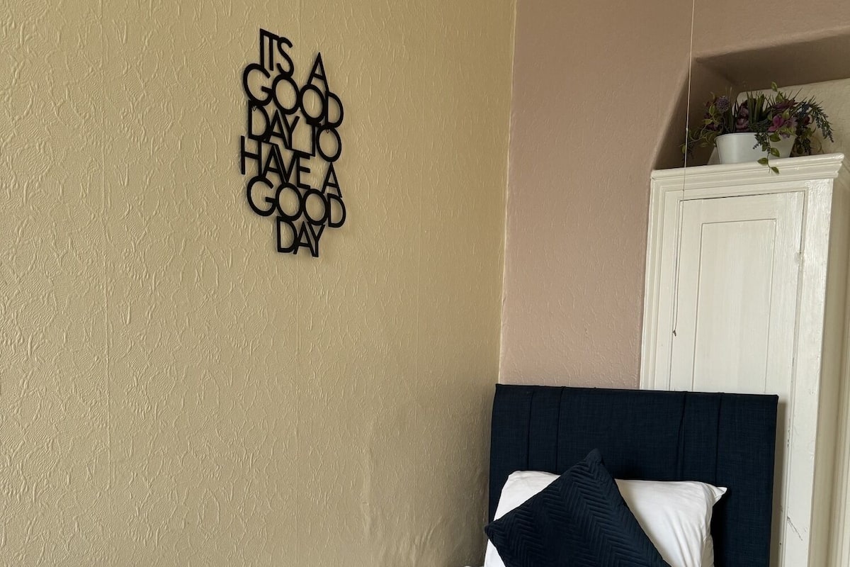 Cama bem arrumada com roupa de cama branca e azul-marinho, um objeto decorativo na parede e um armário branco com uma planta.