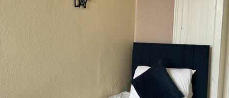 A neatly made bed with white and navy bedding, a decorative wall piece, and a white cabinet with a plant.