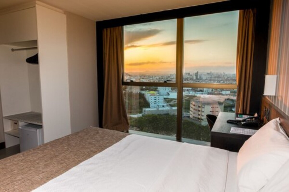 Een hotelkamer met een bed, een bureau, een stoel en een groot raam met uitzicht over de stad.