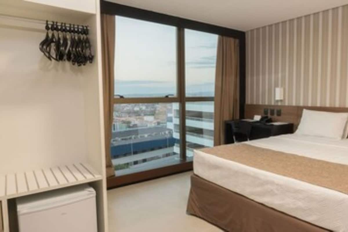 A hotel room with a large bed, a desk, a chair, a wardrobe, and a view of the city through a large window.