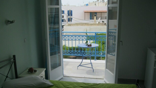 A room with a bed, a small table with a plant, and a balcony with a blue railing and a table. - Very-Kokkos Pension 2 (Naxos)