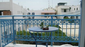 A balcony with a blue metal railing, a small round table with a potted plant, and a view of white buildings in the background. - Very-Kokkos Pension 2 (Naxos)