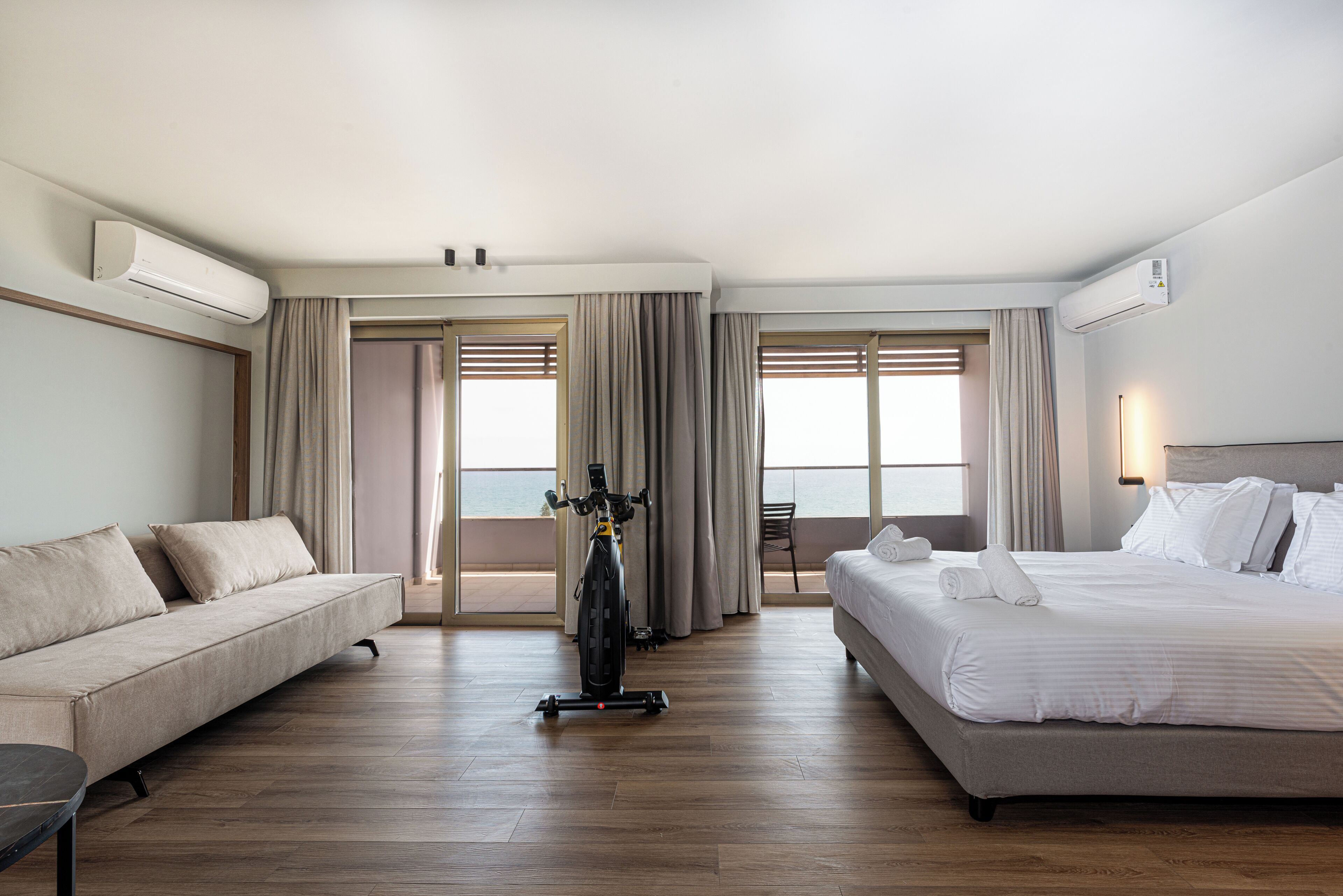 Quarto de hotel com cama, sofá, bicicleta e vista para o mar.