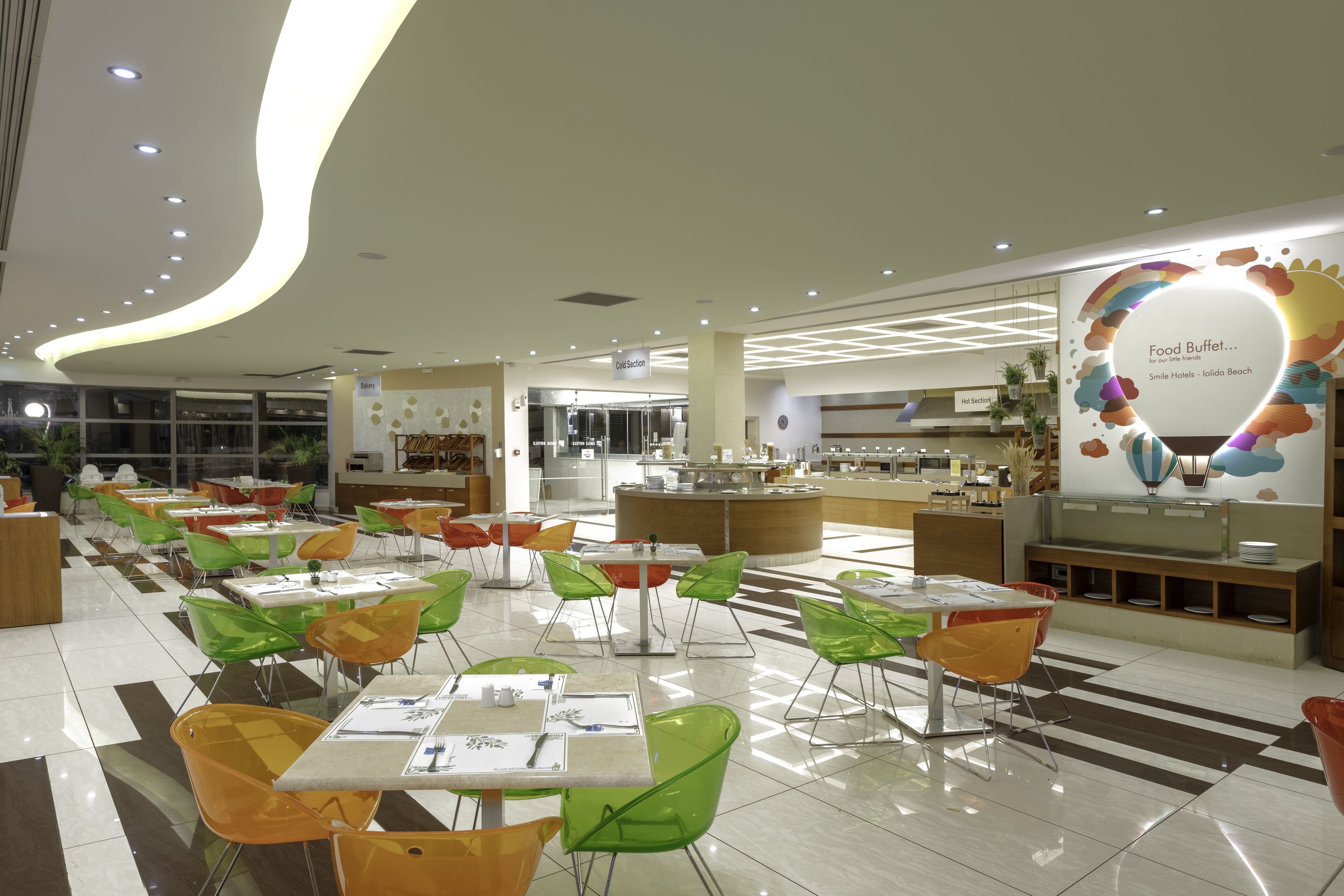 Un comedor moderno con sillas verdes y anaranjadas, un bufé central de comida y un cartel que dice 'Smile Hotels - Ildora Beach'.