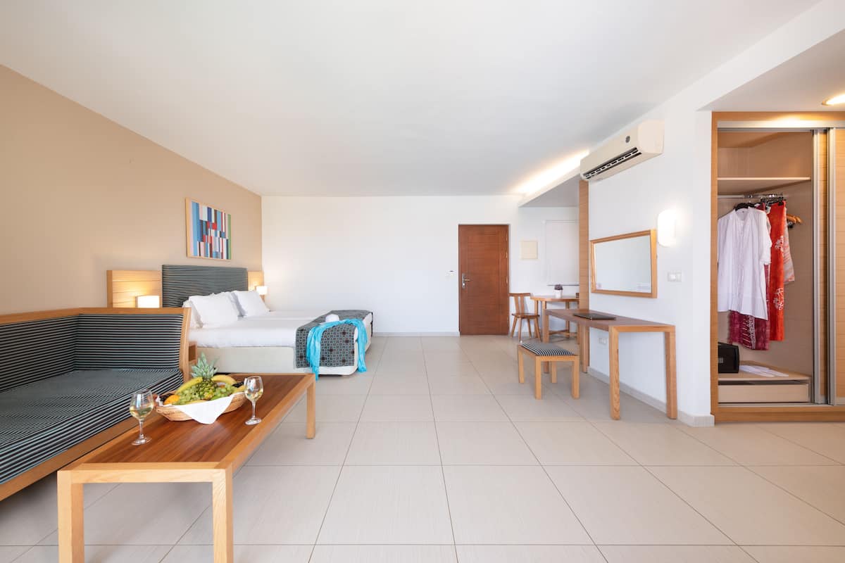 A modern hotel room with a sofa, a dining table, a small kitchen area, and a closet with hanging clothes.